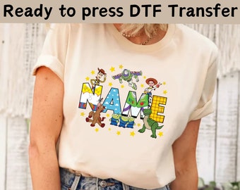 May include: A cream-colored t-shirt with a colorful graphic of characters from the Toy Story movie franchise. The graphic includes Woody, Buzz Lightyear, Jessie, and other characters. The word "NAME" is spelled out in large letters with the characters standing on top of the letters. The graphic is surrounded by yellow stars.