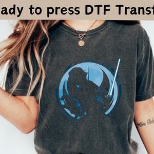 以下が含まれることがあります： 青いグラフィックが特徴の濃い灰色のTシャツ。ライトセーバーを持った人物が円形デザインの中に描かれています。「Ready to press DTF Transfer」の文字が上部にあります。Tシャツは人が着ています。