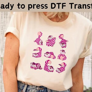 Puede incluir: Una camiseta blanca con un diseño de Gato de Cheshire rosa y blanco. El diseño presenta al Gato de Cheshire en varias poses, incluyendo sonriendo, sonriendo y estirándose. El texto "Ready to press DTF Transfer" está en la parte superior de la imagen.