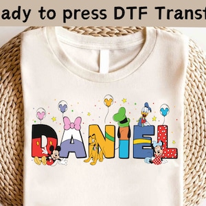Op de afbeelding: Crèmekleurig T-shirt met de naam "DANIEL" in kleurrijke, cartoonachtige letters. Elke letter heeft een ander Disney-personage, waaronder Mickey Mouse, Minnie Mouse, Pluto en Donald Duck. De tekst "Ready to press DTF Transfer" staat bovenaan.
