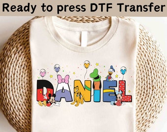 May include: Cream-colored t-shirt with the name "DANIEL" in colorful, cartoon-style letters. Each letter features a different Disney character, including Mickey Mouse, Minnie Mouse, Pluto, and Donald Duck. The text "Ready to press DTF Transfer" is at the top.