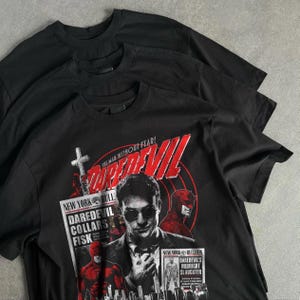 May include: Three black t-shirts are stacked, with the front shirt featuring a graphic of the Daredevil comic book character. The graphic includes the text "DAREDEVIL" in red, the phrase "THE MAN WITHOUT FEAR!", and other text related to the comic.