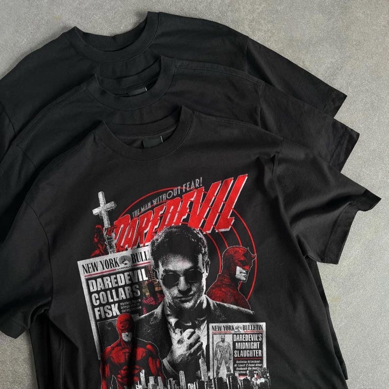May include: Three black t-shirts are stacked, with the front shirt featuring a graphic of the Daredevil comic book character. The graphic includes the text "DAREDEVIL" in red, the phrase "THE MAN WITHOUT FEAR!", and other text related to the comic.