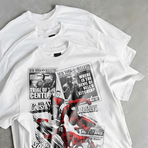 May include: Three white crew neck t-shirts are displayed. The bottom shirt has a comic book-style design with the text "NEW YORK BULLETIN," "TRIAL OF THE CENTURY," "DAREDEVIL," and "SPIDER-MAN." The graphic is in black and red.