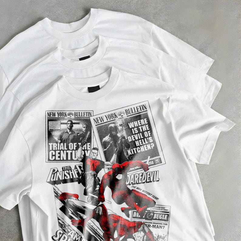 May include: Three white crew neck t-shirts are displayed. The bottom shirt has a comic book-style design with the text "NEW YORK BULLETIN," "TRIAL OF THE CENTURY," "DAREDEVIL," and "SPIDER-MAN." The graphic is in black and red.
