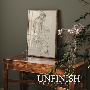 May include: A black and white line drawing of a person sitting, framed in a light brown wood frame, on a wooden table with a vase of white flowers.