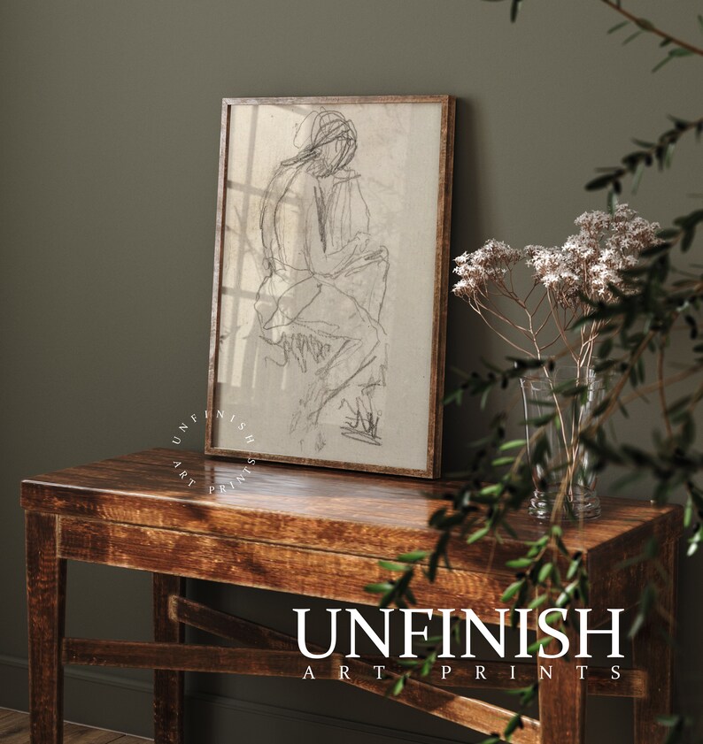 May include: A black and white line drawing of a person sitting, framed in a light brown wood frame, on a wooden table with a vase of white flowers.