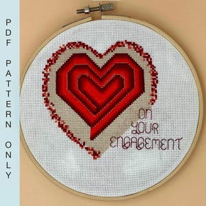 Könnte beinhalten: Eine Kreuzstich-Stickerei in einem Holzrahmen zeigt ein geschichtetes rotes Herzdesign. Die Worte "ON YOUR ENGAGEMENT" sind darunter gestickt. Der Text "PDF PATTERN ONLY" befindet sich auf der linken Seite.