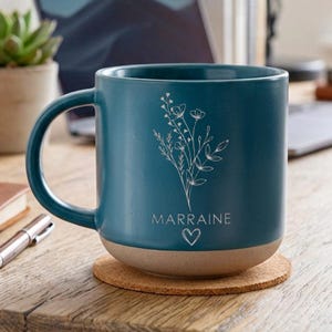 May include: A teal ceramic mug with a white floral design and the word "MARRAINE" with a heart symbol. The bottom of the mug is a tan color. The mug is sitting on a round cork coaster.