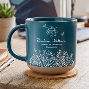 May include: A teal ceramic mug with a brown base, personalized with the name "Sophia Miller", Cambridge University, and "Class of 2026". The mug features a floral design and graduation-themed illustrations.