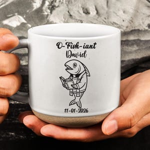 May include: A white ceramic mug with a speckled base featuring a black line drawing of a fish dressed as a clergy member holding a book. The text reads O-Fish-iant David above the fish and 11-01-2026 below the illustration.