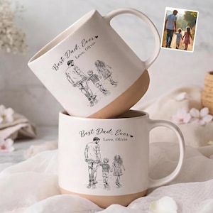 May include: Two white ceramic mugs with a tan base, stacked. Each mug features a black sketch of a father walking with two children, with the text "Best Dad, Ever" and "Love, Olivia". The mugs have large, curved handles.