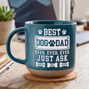 May include: A teal ceramic mug with the text "BEST DOG DAD EVER, EVER, EVER JUST ASK" in white. The mug has a brown base and the names "CHARLIE", "MAX", and "COOPER" below the text. The mug is on a wooden surface.