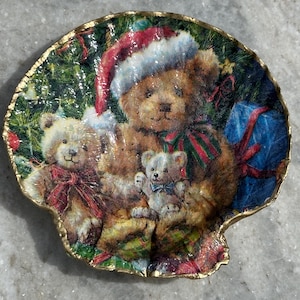 Puede incluir: Un plato decorativo con forma de concha marina con un diseño navideño. El plato muestra tres osos de peluche, uno con un gorro de Papá Noel, rodeados de regalos y vegetación festiva. El plato tiene un borde dorado.