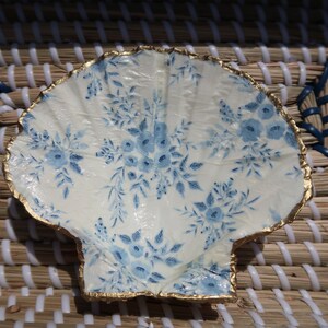 May include: A white seashell-shaped dish with a blue and white floral pattern and a gold rim.