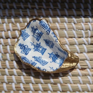 May include: A white ceramic oyster shell dish with a blue and white pattern of pagodas. The dish is painted with gold around the edges.