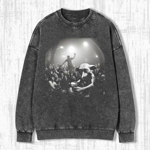 May include: A black and white graphic sweatshirt with a crew neck and long sleeves. The graphic depicts a crowd scene with a creature holding a cup. The sweatshirt has a faded, distressed appearance.