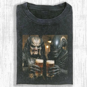 May include: Dark grey t-shirt with a graphic of a Predator and an Alien raising beer glasses. The image is centrally placed, showing the creatures holding the beer. The shirt has a crew neck and a slightly faded, vintage look.