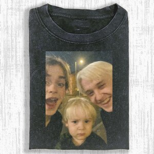 May include: Dark gray t-shirt featuring a faded photograph of three people. The photograph shows two adults and a child, all smiling or with surprised expressions. The t-shirt has a vintage, worn look.