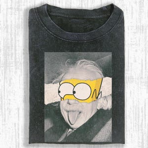 May include: Dark gray t-shirt featuring a graphic of Albert Einstein with a yellow cartoon eye mask. The mask covers the eyes and is torn to reveal the eyes. The shirt is a crew neck.