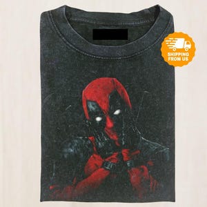 May include: A folded, dark gray t-shirt featuring a graphic of Deadpool in his signature red and black costume. The image shows Deadpool with his hands near his face, with glowing white eyes. The shirt has a vintage, textured appearance.