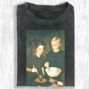 May include: Dark gray t-shirt featuring a vintage-style image of two people cooking in a kitchen. The image has a faded, retro aesthetic. One person stirs a pan with a wooden spoon, while the other whisks in a bowl. The shirt is folded.