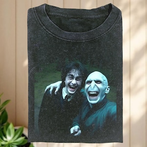 Könnte beinhalten: Dunkelgraues T-Shirt mit einer Grafik von zwei lachenden Charakteren aus der Harry-Potter-Reihe. Eine Figur trägt eine Brille und eine Krawatte, die andere hat ein blasses Gesicht und trägt eine dunkelgrüne Robe. Das Bild ist vor einem grünen Hintergrund.