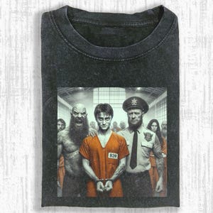 May include: Dark gray t-shirt featuring a graphic of a person in an orange jumpsuit with handcuffs, flanked by a guard and a creature. The image has a prison theme. The shirt is made of a soft material.