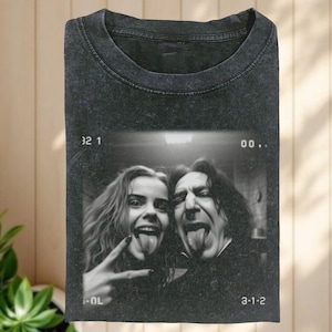 Puede incluir: Camiseta gris oscuro con una foto en blanco y negro de dos personas sacando la lengua. La imagen tiene un borde de película retro con números. La camiseta está doblada y tiene cuello redondo.