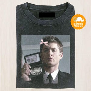 May include: Dark gray t-shirt featuring a graphic of a man holding a badge, with a pink bow on his head. The image is printed on a pre-washed, vintage-style tee.