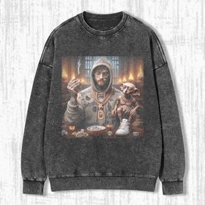 May include: Dark gray sweatshirt with a graphic print. The print features a person in a hoodie with gold jewelry, smoking a cigarette, and a small creature. The sweatshirt has a crew neck and long sleeves.