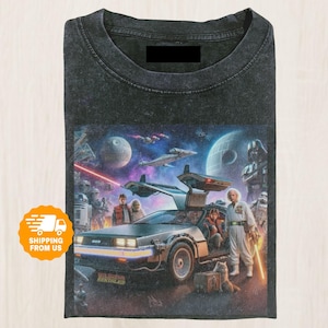 May include: Dark gray t-shirt featuring a colorful graphic with characters and vehicles from the Back to the Future and Star Wars franchises. The graphic includes the DeLorean time machine, spaceships, and iconic characters.