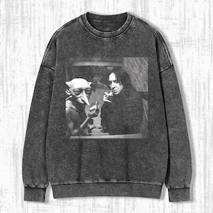 Peut inclure: Un sweat-shirt gris anthracite avec une image en noir et blanc de deux personnages. L'un est un homme grand aux cheveux noirs, l'autre une créature avec de grandes oreilles et un long nez. Ils fument tous les deux des cigarettes.