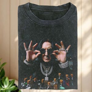 May include: Dark gray t-shirt featuring a portrait of a man wearing round sunglasses and multiple rings. The man is surrounded by bottles with cork stoppers. The shirt has a vintage, washed look.