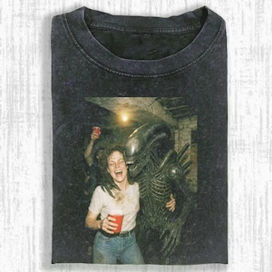 May include: Dark grey t-shirt featuring a vintage-style image of a woman laughing while embracing an alien. The woman is holding a red cup. The alien is dark green. The shirt has a crew neck.