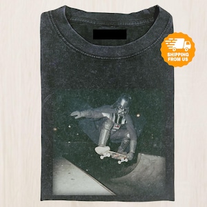 Peut inclure: T-shirt gris foncé avec un motif de Dark Vador faisant du skateboard. L'image montre Dark Vador dans son casque et sa combinaison, en l'air sur un skateboard dans un skate park. Le t-shirt a un col rond et un aspect légèrement délavé.