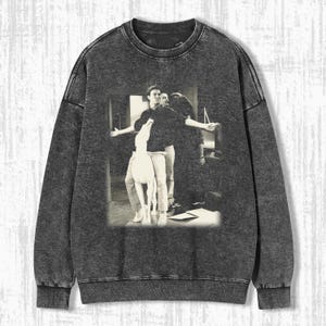 以下が含まれることがあります： テレビ番組Friendsのシーンをあしらった、色あせた白黒グラフィックのダークグレーのスウェットシャツ。グラフィックは、腕を広げた3人の人物が部屋にいる様子を描いています。スウェットシャツはクルーネックと長袖です。