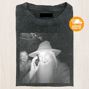 以下が含まれることがあります： 電話をしている魔法使いの白黒画像がプリントされた黒いTシャツ。魔法使いは帽子をかぶり、長い白いひげを生やしています。背景には別の人物がいます。Tシャツは折りたたまれています。