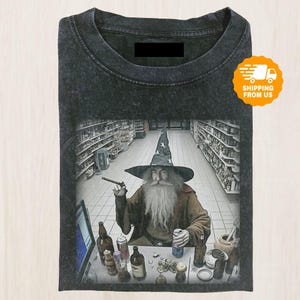 May include: Dark gray t-shirt featuring a wizard in a hat smoking a cigar, seated at a table with bottles and cans. The wizard is in a grocery store setting. The shirt has a vintage, washed look.