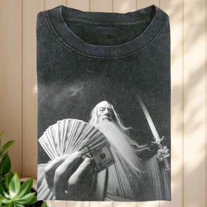 Peut inclure: T-shirt gris foncé avec un motif en noir et blanc d'un magicien tenant un éventail de billets de 100 dollars américains et une épée. Le magicien a une longue barbe blanche et porte une robe.