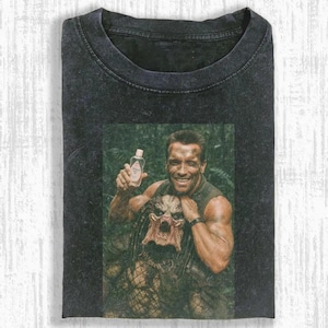 May include: Dark grey t-shirt with a humorous image of a smiling man holding a baby lotion bottle, with a Predator creature. The image is set against a jungle backdrop. The shirt is folded.