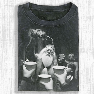 May include: Dark gray t-shirt featuring a black and white graphic of a wizard pouring from a bottle into cups. The image has a vintage, distressed look, with a crew neck and short sleeves.