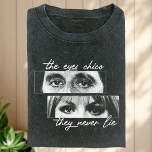 May include: Dark grey t-shirt with the text "the eyes chico they never lie" in white script. The shirt features a graphic of two pairs of eyes in rectangular boxes, one above the other.