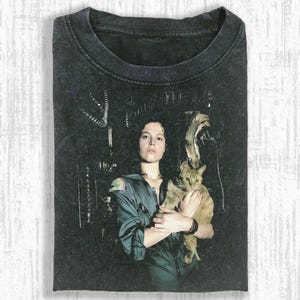 May include: Dark gray t-shirt featuring a portrait of a person holding a cat. The person is wearing a green jumpsuit. The background has a dark, industrial aesthetic. The cat is light brown with dark stripes.