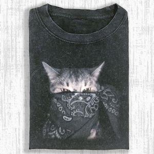 Puede incluir: Camiseta negra con un gato que lleva un pañuelo negro. Los ojos del gato son visibles por encima del pañuelo. La camiseta está doblada y tiene un aspecto vintage y desgastado.
