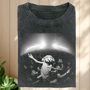 Puede incluir: Camiseta gris oscuro con una imagen en blanco y negro de una criatura con orejas grandes y una multitud de personas. La criatura está en el centro con los brazos extendidos. La camiseta tiene cuello redondo.