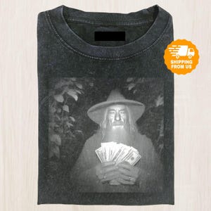 May include: Dark gray t-shirt featuring a black and white image of a man in a hat holding money. The image is centered on the shirt. The shirt has a crew neck and short sleeves. The shirt is folded.