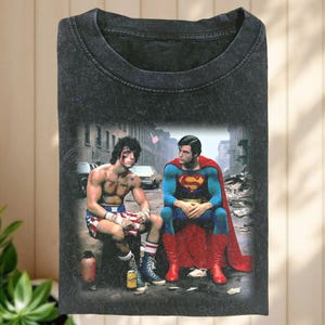 Könnte beinhalten: Dunkelgraues T-Shirt mit einem Aufdruck von Rocky Balboa und Superman, die zusammen sitzen. Rocky trägt Boxershorts und Superman sein ikonisches Kostüm. Das Bild vor einem Stadtpanorama.