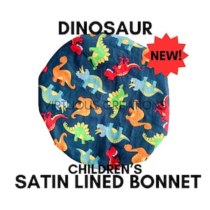 May include: A navy blue satin-lined bonnet for children, patterned with colorful cartoon dinosaurs in shades of orange, green, red, and blue. The word "DINOSAUR" is printed above the bonnet, and "NEW!" is in a red starburst.