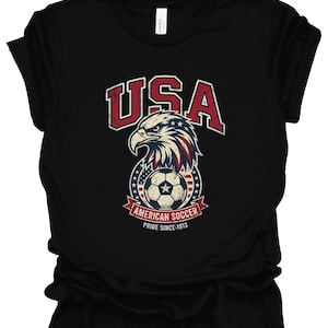 K&ouml;nnte beinhalten: Ein schwarzes Kurzarm-T-Shirt mit einer Grafik eines Wei&szlig;kopfseeadler-Kopfes &uuml;ber einem Fu&szlig;ball, mit dem Text USA, AMERICAN SOCCER und PRIDE SINCE 1913. Das Design enth&auml;lt rote, wei&szlig;e und blaue Farben mit einer abgenutzten, vintage-artigen Textur.